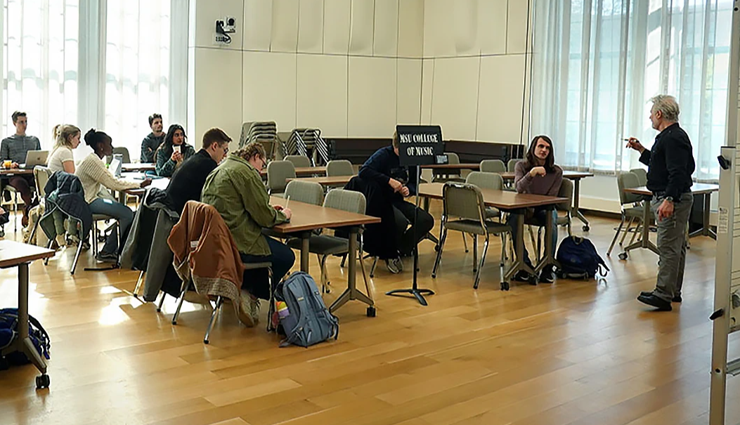 A large classroom with tall windows and wood floors is filled with several desk. Students can be seen seated at the desks with their backpacks, they are looking to the front of the room where a man stands addressing them.